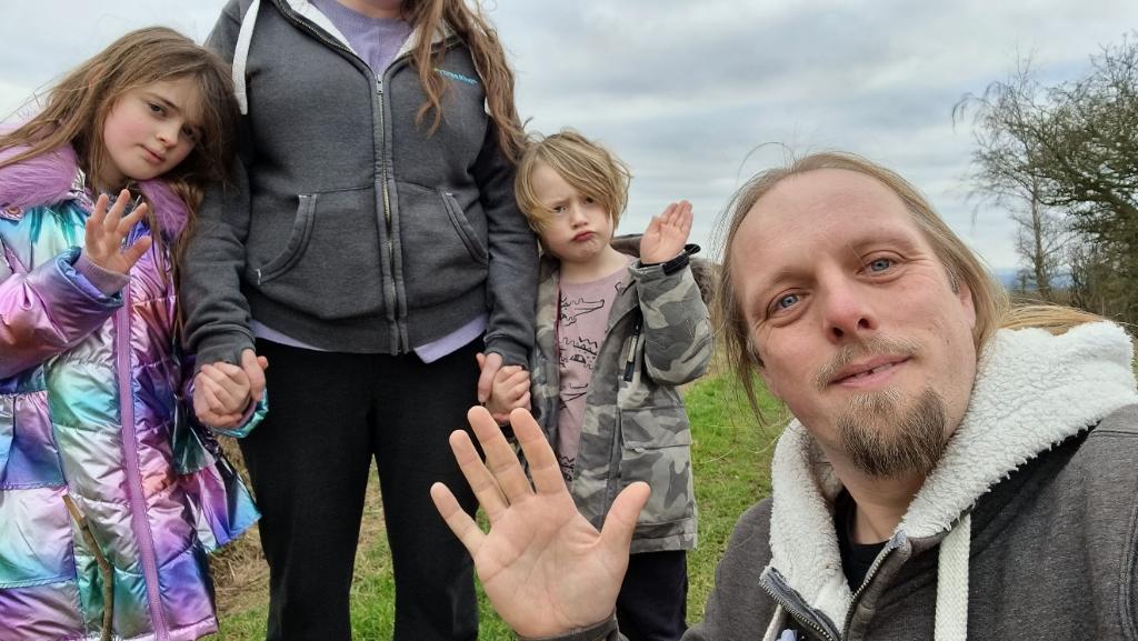 Dan smiles and waves from a grassy hilltop; behind him, two children - a girl and a boy - wave too, but half-heartedly and with tired expressions. The sky behind is grey and cloudy.