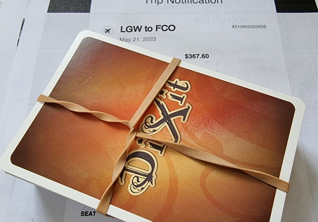 A deck of Dixit cards, bound by a twisted elastic band, sits on a flight itinerary for the journey "LGW to FCO" taking place on May 21, 2023 and costing $367.60.