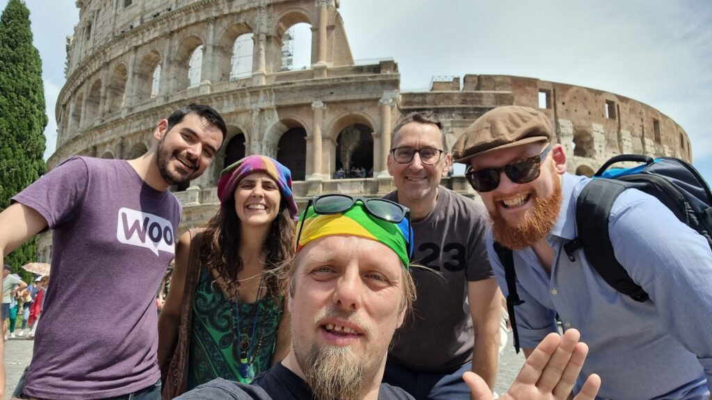 In front of the Colosseum in Rome, Dan - wearing a rainbow-striped bandana atop which his sunglasses are perched - takes a selfie. Behind him stand a man with dark hair and a closely-trimmed beard wearing a purple "woo" t-shirt, a woman with long brown hair wearing beads and a multicoloured dress, a man wearing spectacles and a dark t-shirt on which the number "23" can be made out, and a man in sunglasses with a ginger beard, wearing an open blue shirt.