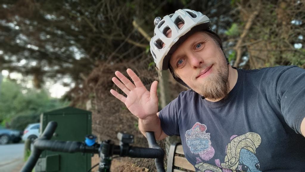 Dan, wearing a white cycle helmet and a worn block t-shirt, waves to the camera while sitting on a bench. Alongside him can be seen the racing/road-bike style handlebars of a bike.