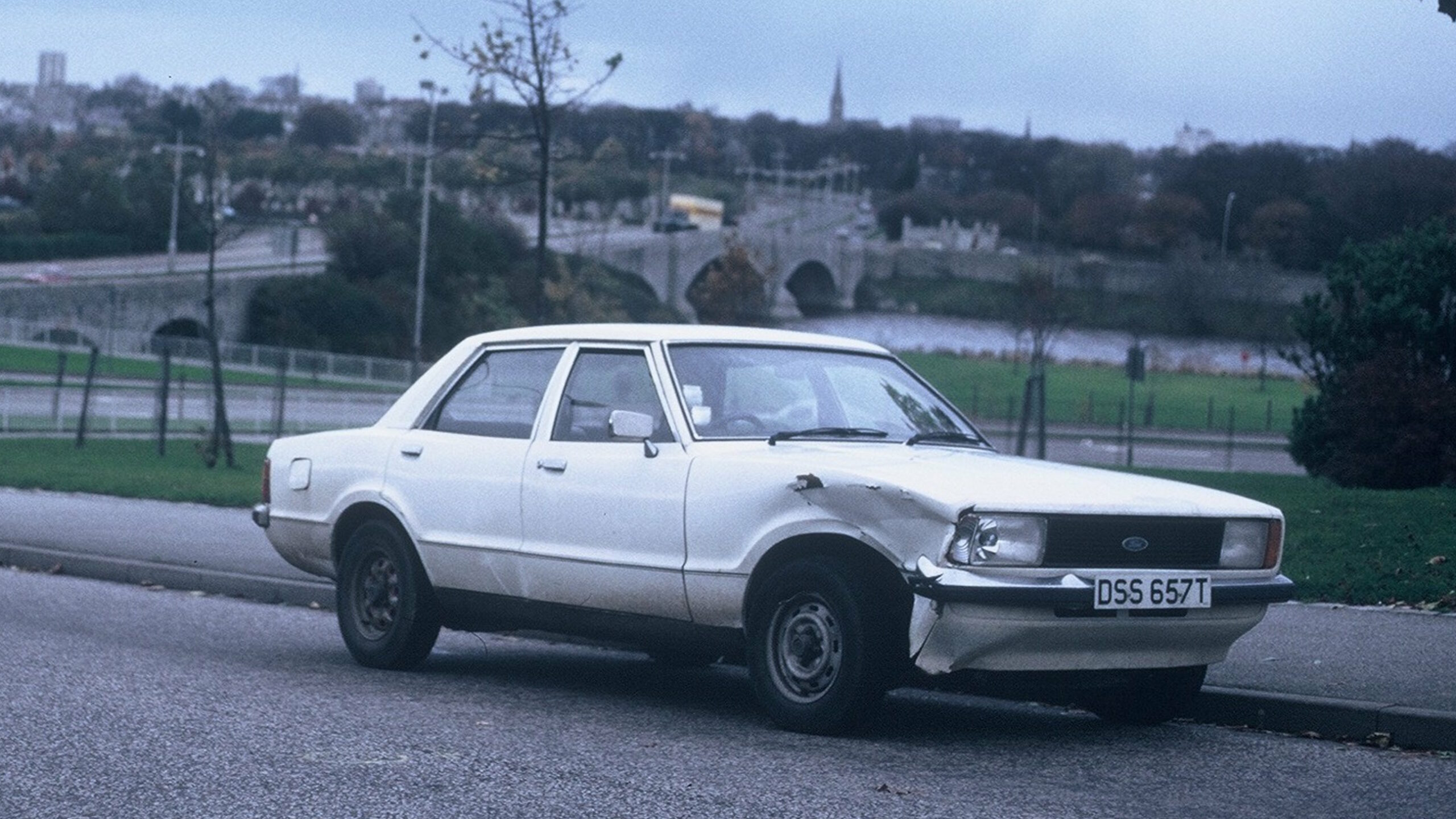 A boxy 1979 white Ford car, number plate DSS 657T with a badly dented and somewhat corroded front wheel arch on the drivers' side, sits empty and parked at the side of an otherwise empty asphalt strreet. In the background, under grey skies, a city skyline can be made out with houses, tower blocks, and a church steeple, on the other side of an arched river bridge. The leaves are early-autumn coloured: mostly greem, but with some brown appearing and a handful of bare branches exposed.