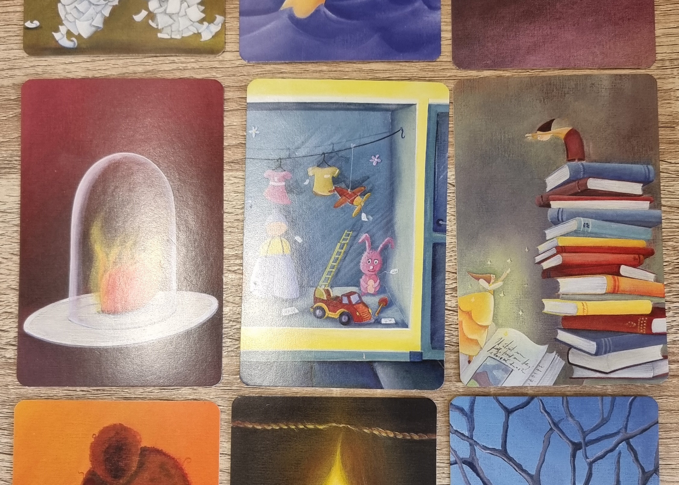 A trio of Dixit cards within a grid of nine. From left to right, they show: a heart, on fire, beneath a glass jar; a cubbyhole containing childrens' toys; a fairy leaping from a book towards a small person atop a stack of books.