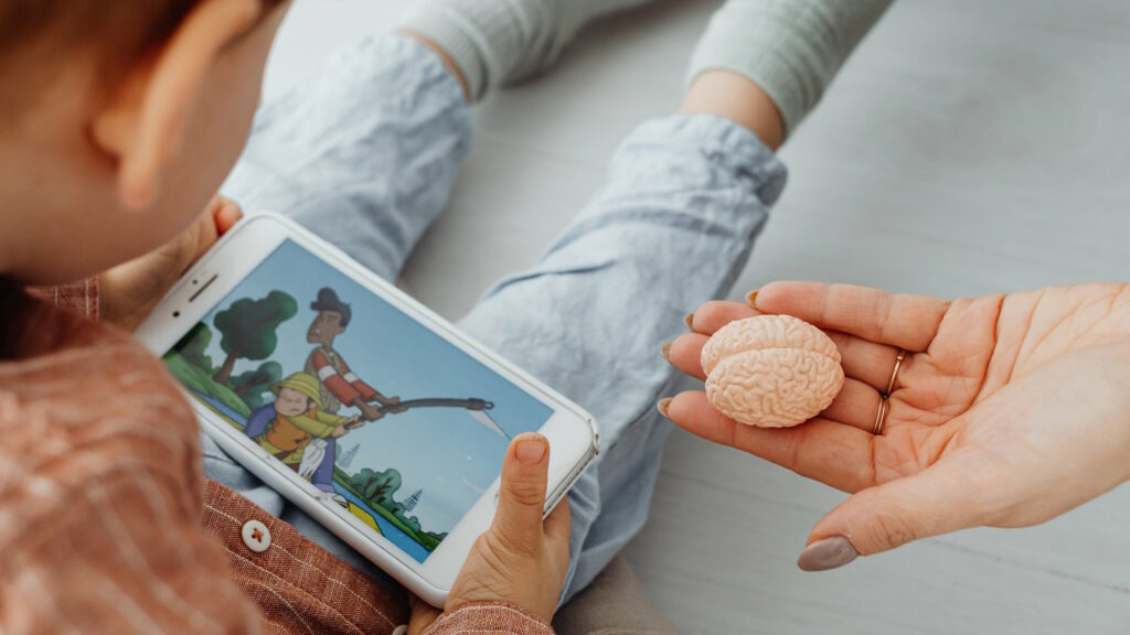 A small child, sitting on the floor, uses a mobile phone to watch a cartoon of two people struggling to pull a fishing rod. A feminine hand with brown-painted nails and rings on two fingers reaches in to offer the child a minature model of a human brain.