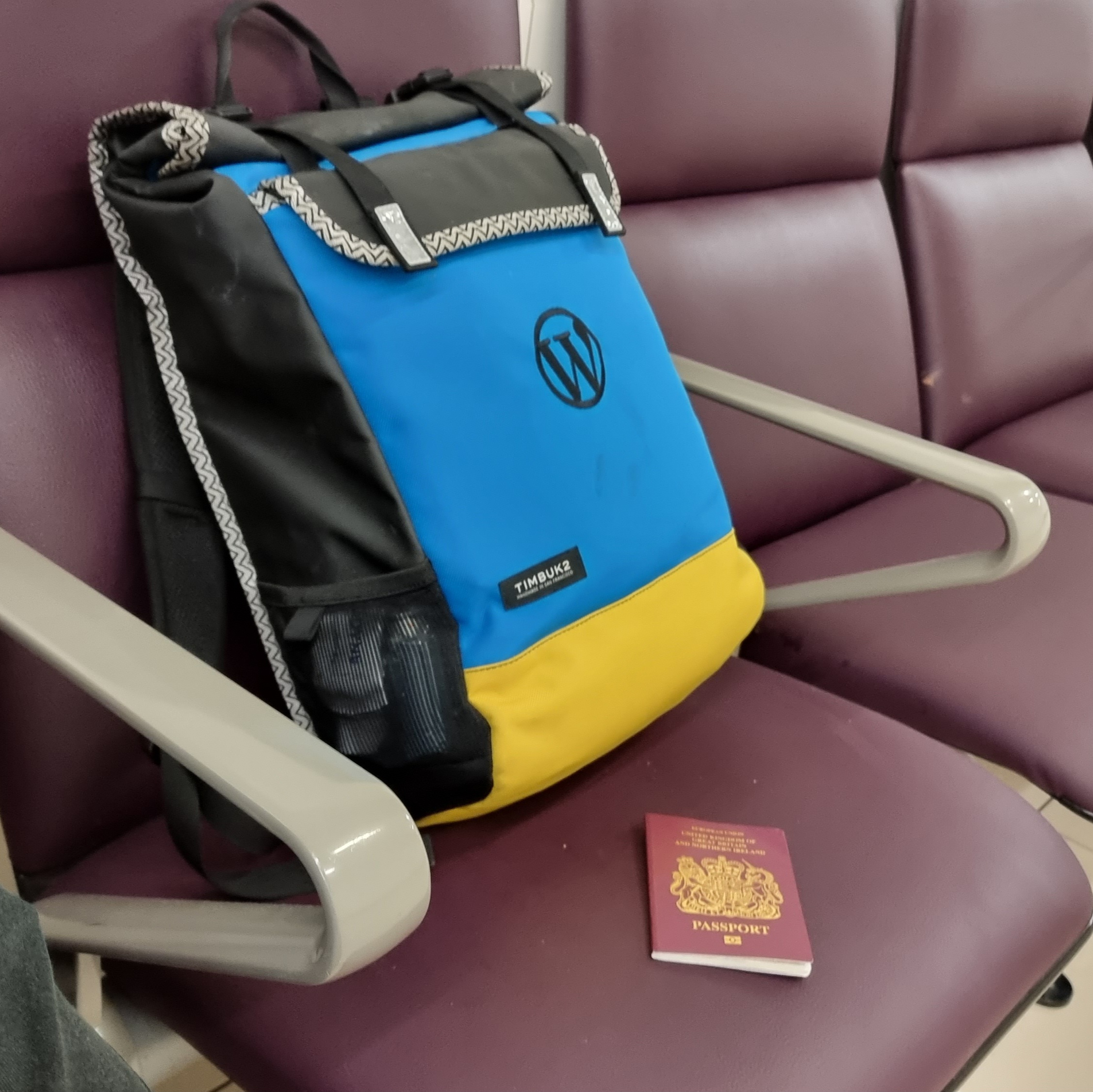 A modest-sized backpack in blue and yellow, with a WordPress logo stiched on, sits on an airport departure lounge bench. Alongside it is a burgundy-coloured British passport.
