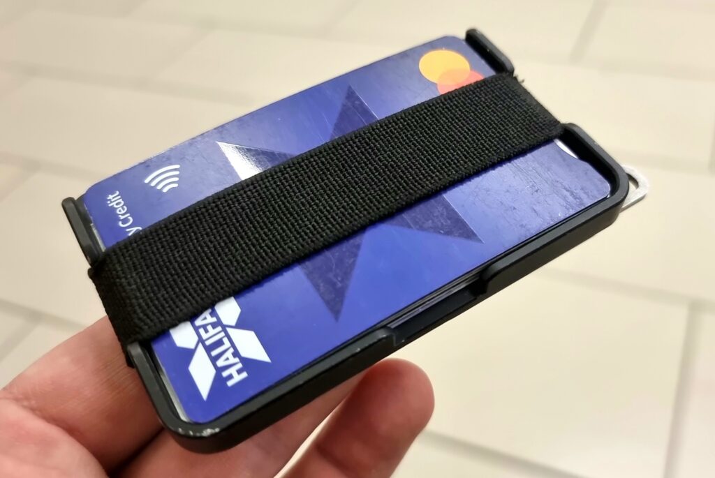 Minimalist carbon fibre wallet, balanced on two fingertips, with parts of a Halifax Mastercard credit card showing from behind an elasticated band.