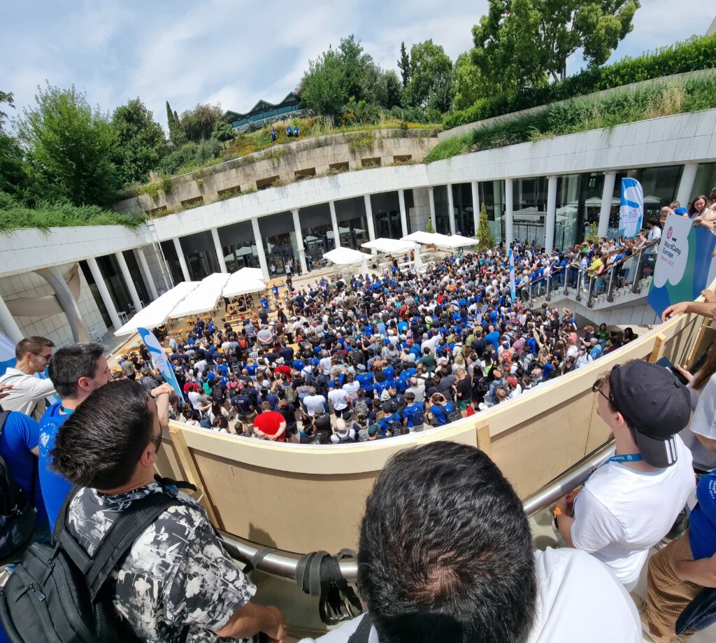An enormous crowd shuffles tightly into a courtyard. A trio of blue-shirted photographers stands atop a building opposite them.