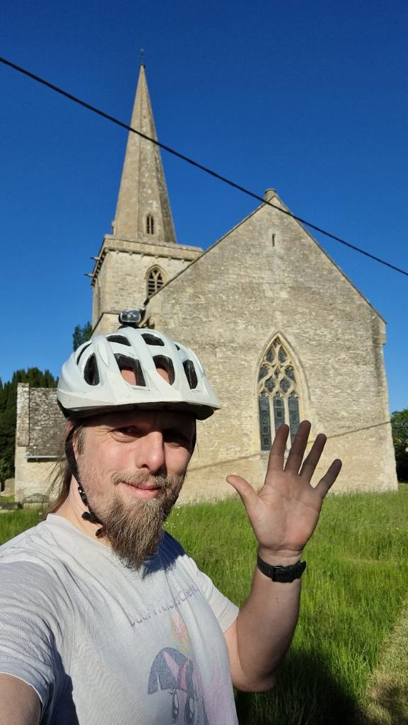 Dan, wearing a cycle helmet, stands in front of a church steeple under a bright blue sky, waving.