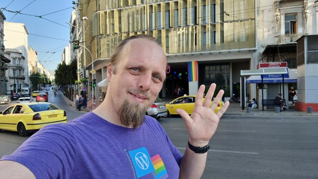 Dan, wearing a purple shirt with a WordPress logo and a pride rainbow, waves to the camera in front of a hotel with pride rainbow banners hanging from its pillars.