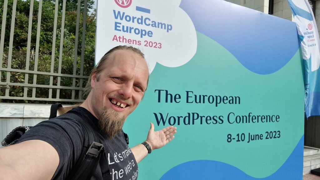 Dan, wearing an Automattic "Let's make the Web a better place" t-shirt, stands in front of a banner welcoming attendees to WordCamp Europe Athens 2023.