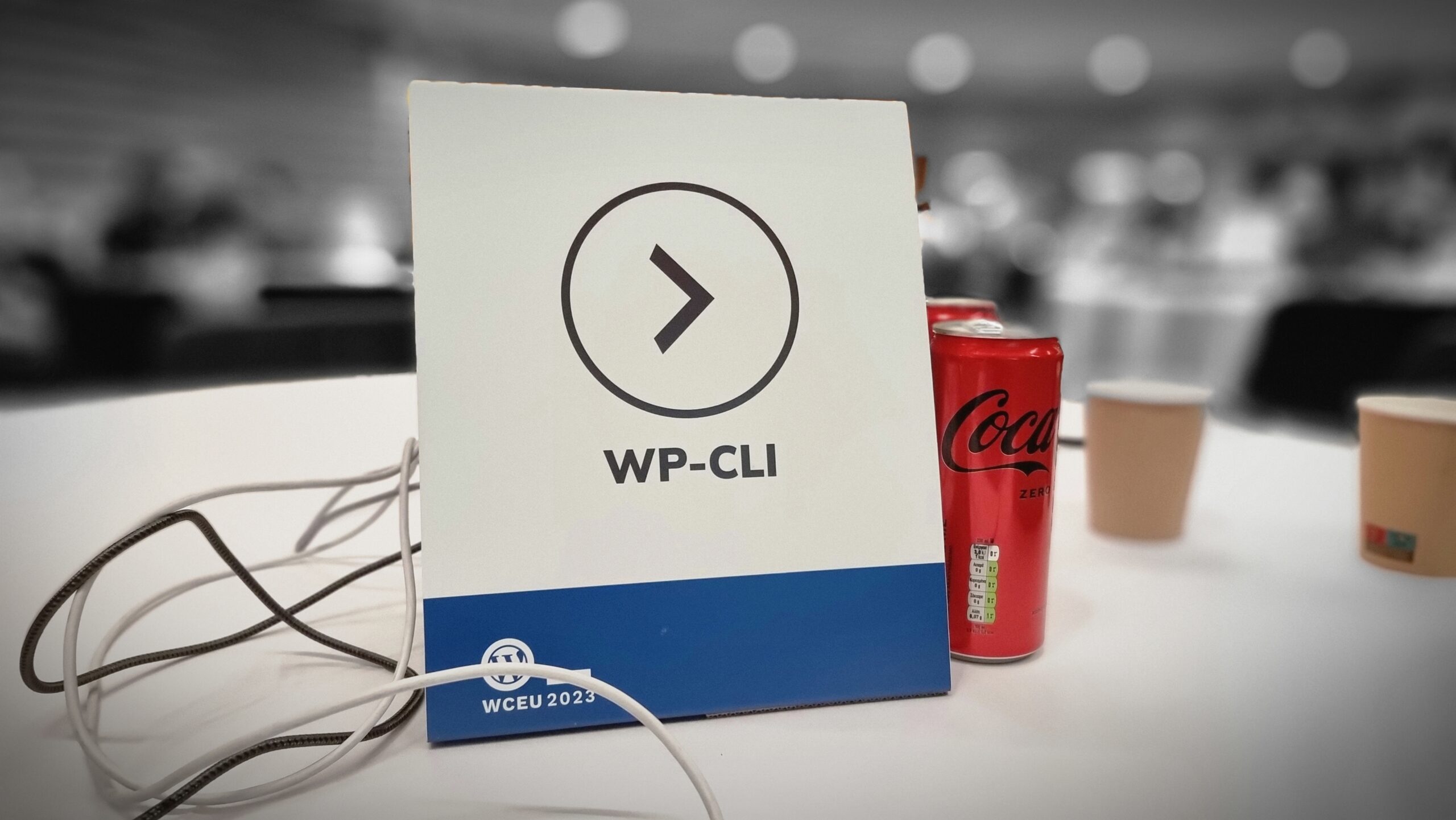 A table placeholder labelled "WP-CLI". It and s handful of Coke cans and disposable coffee cups are picked-out in colour on an otherwise monochrome and blurred picture.