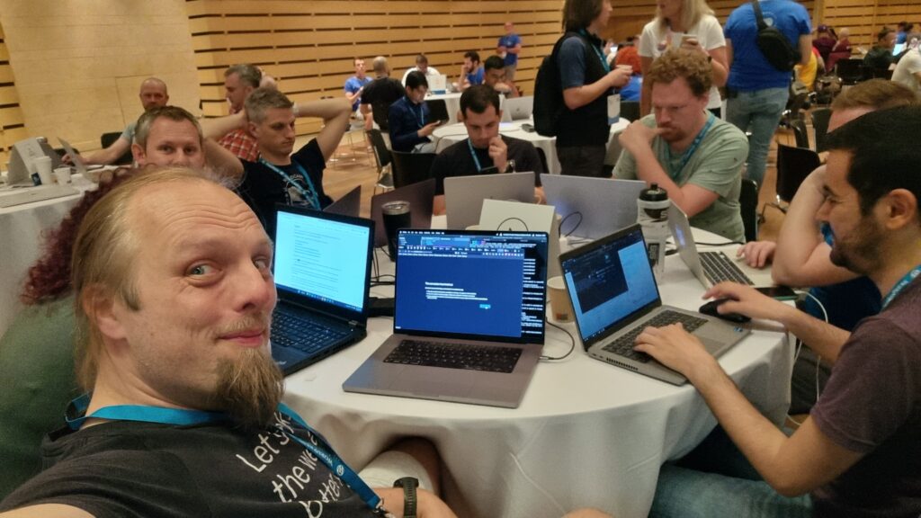 Dan takes a selfie from a round table covered in laptops, with people hacking at them.