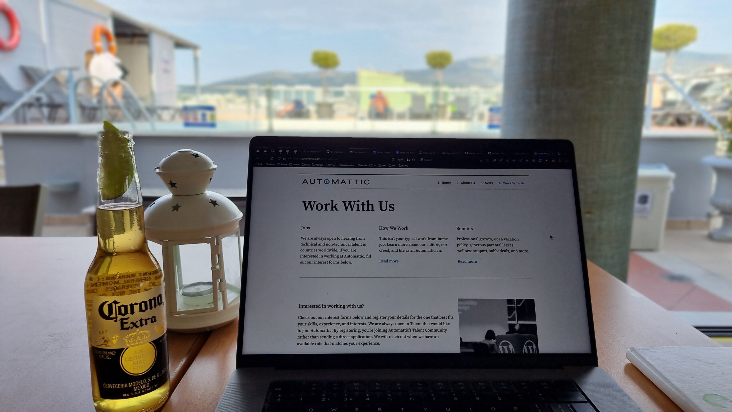 MacBook showing an Automattic "Work For Us" web page, alongside a bottle of Corona Extra. A rooftop terrace garden and swimming pool can be seen in the background.