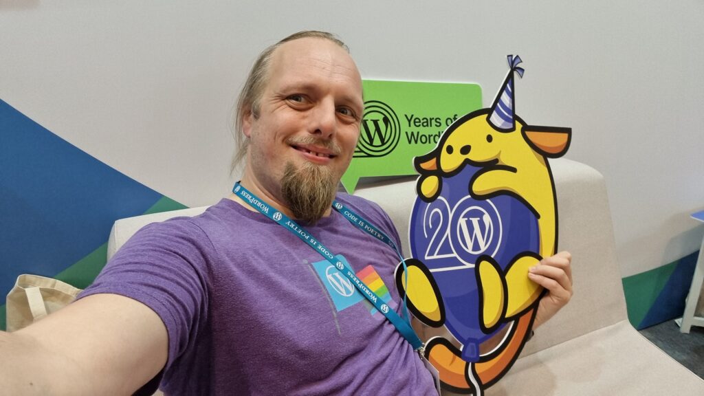 Dan, smiling, wearing a purple t-shirt with a WordPress logo and a Pride flag, hugs a cut-out of a Wappu (itself hugging a "WP 20" balloon and wearing a party hat).