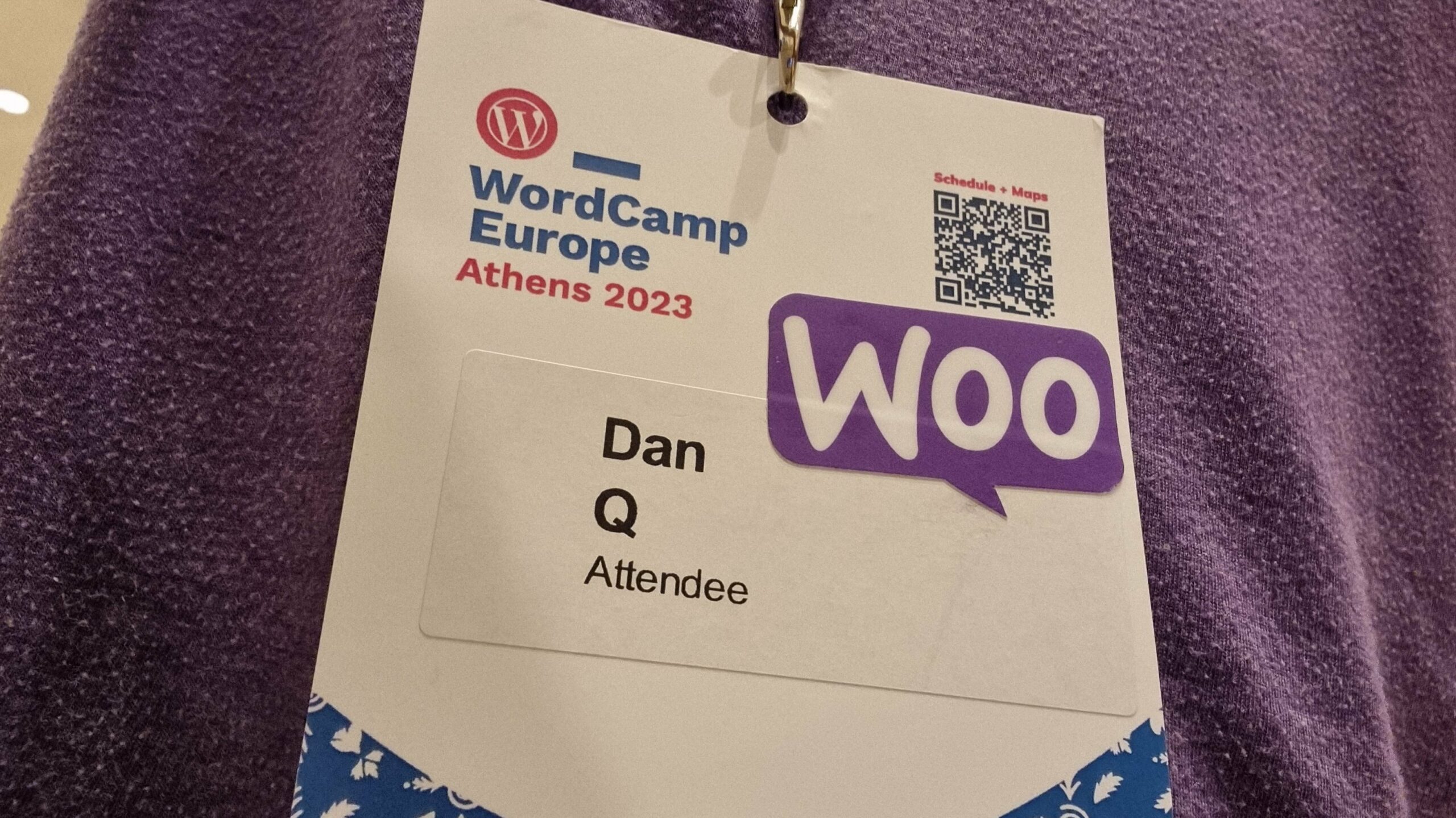 A WordCamp Europe Athens 2023 lanyard and name badge for Dan Q, Attendee, onto which a "Woo" sticker has been affixed.