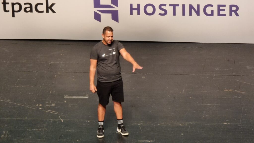 A man on a stage with his arm out in greeting to the crowd in front of him.