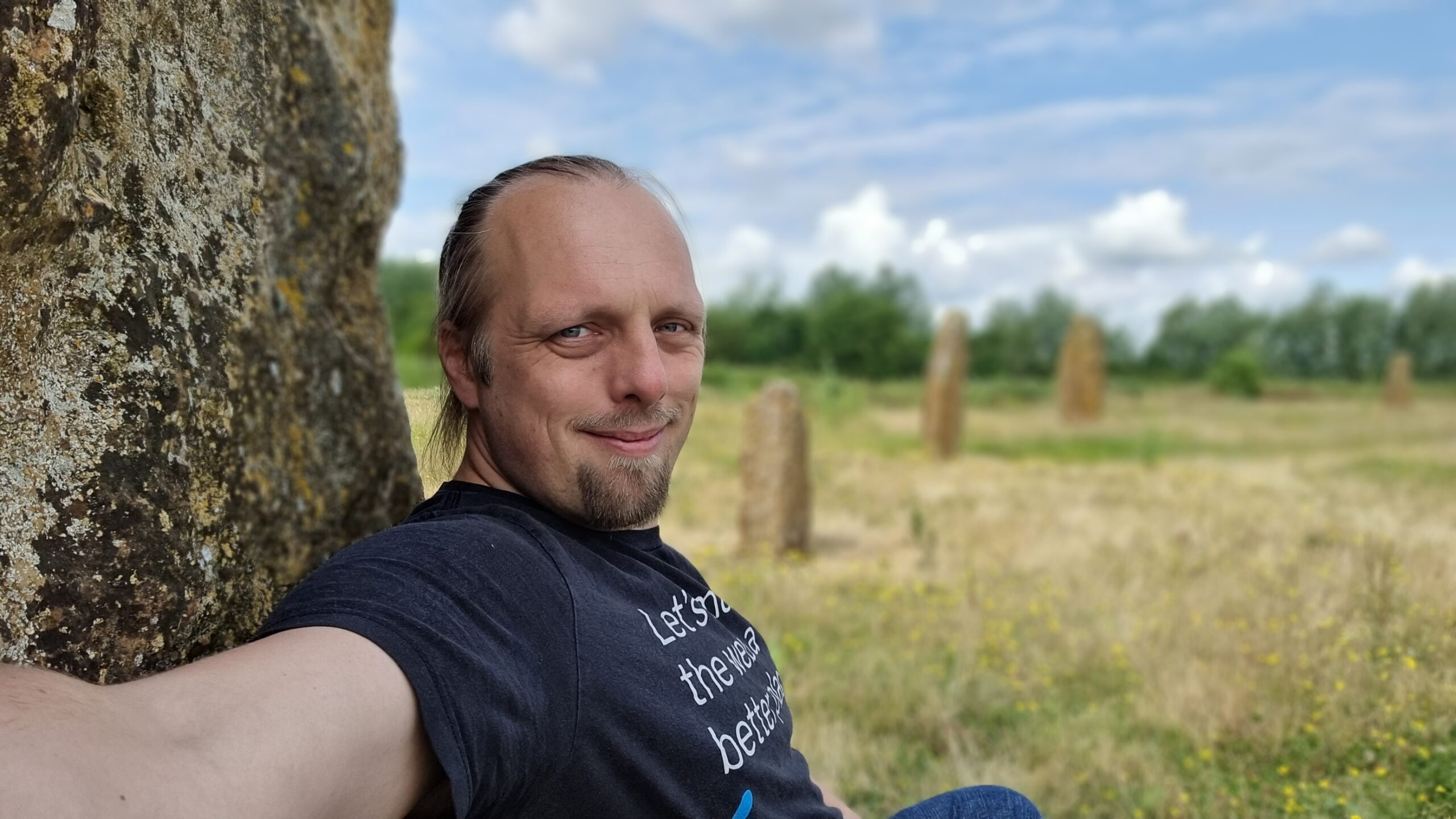 Dan, wearing a black t-shirt with the words "Let's make the web a better place" on, sits with his back to a standing stone. Four more standing stones can be seen stretching away into the bakground, atop a flowery meadow and beneath a slightly cloudy but bright sky.