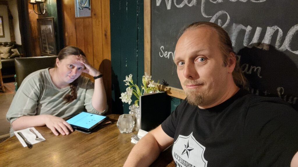 Ruth and Dan, looking tired and frustrated, sit at a pub table. Ruth is using her tablet computer.