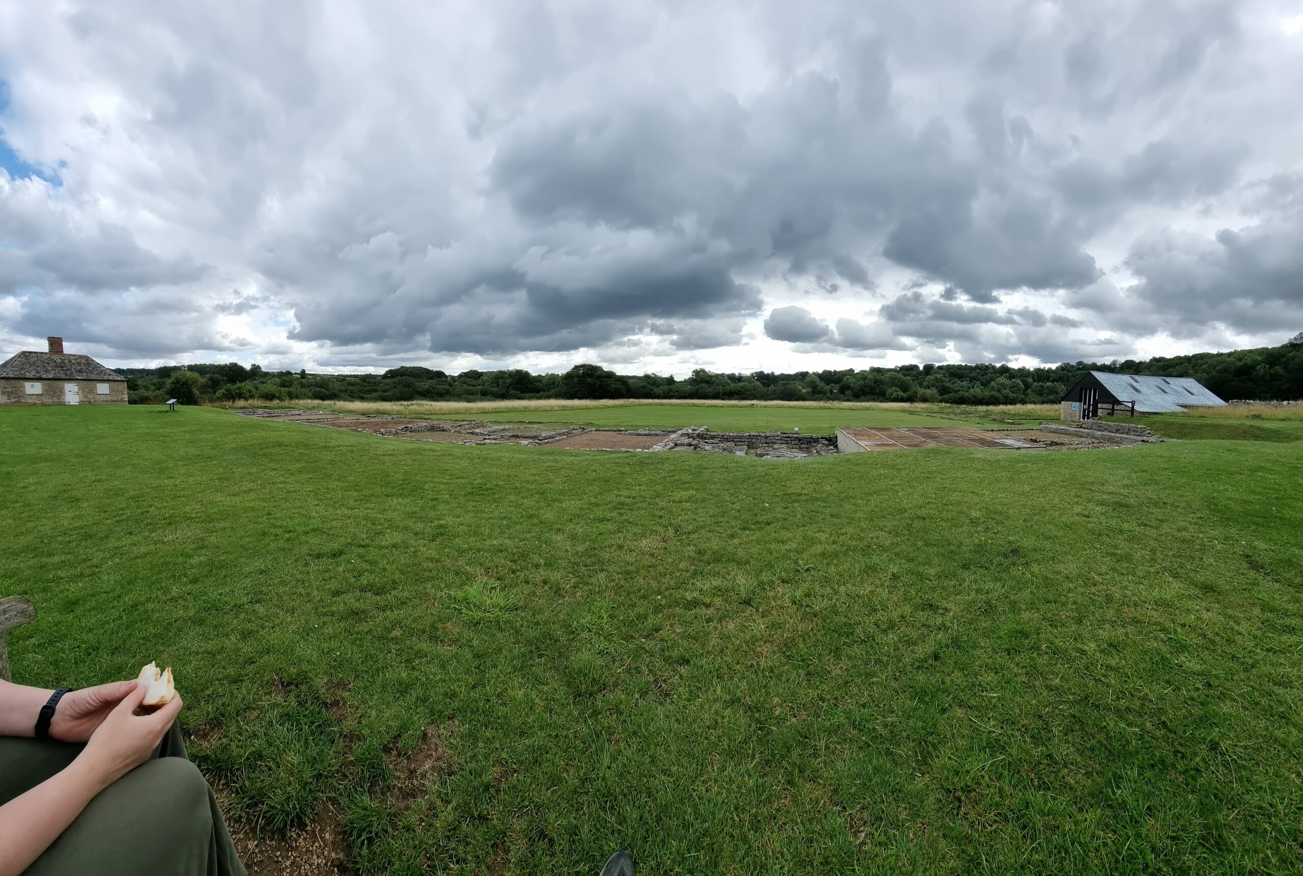 Panoramic photo showing a field containing the remains of a Roman villa in West Oxfordshire, under grey skies. The walls are barely visible in this wide shot.