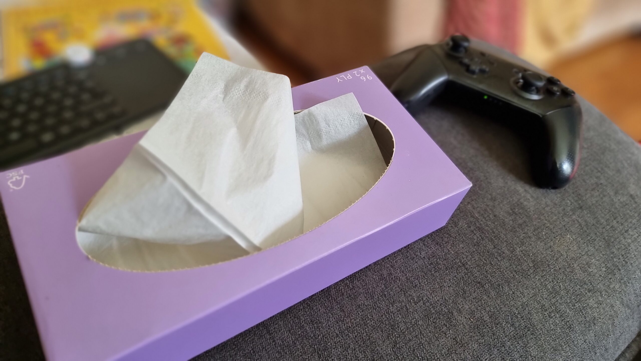 A box of tissues and a Nintendo Switch Pro Controller on the arm of a sofa.