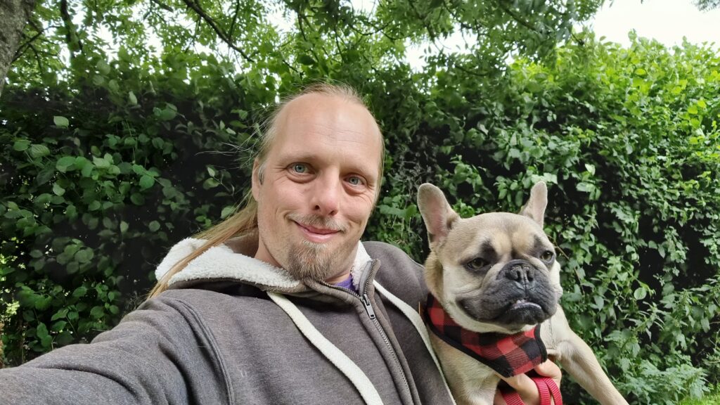 Dan, looking slightly less-unwell, stands holding Demmy, a French Bulldog, in front of a hedge.