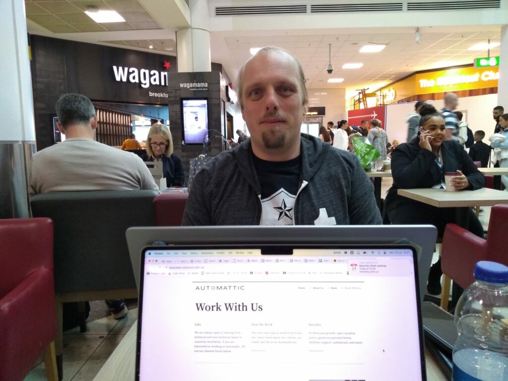 A laptop screen shows Automattic's "Work With Us" web page. Beyond it, in an airport departure lounge (with diners of Wagamama and The Breakfast Club in the background), Dan sits at another laptop, wearing a black "Accessibility Woke Platoon" t-shirt and grey Tumblr hoodie.