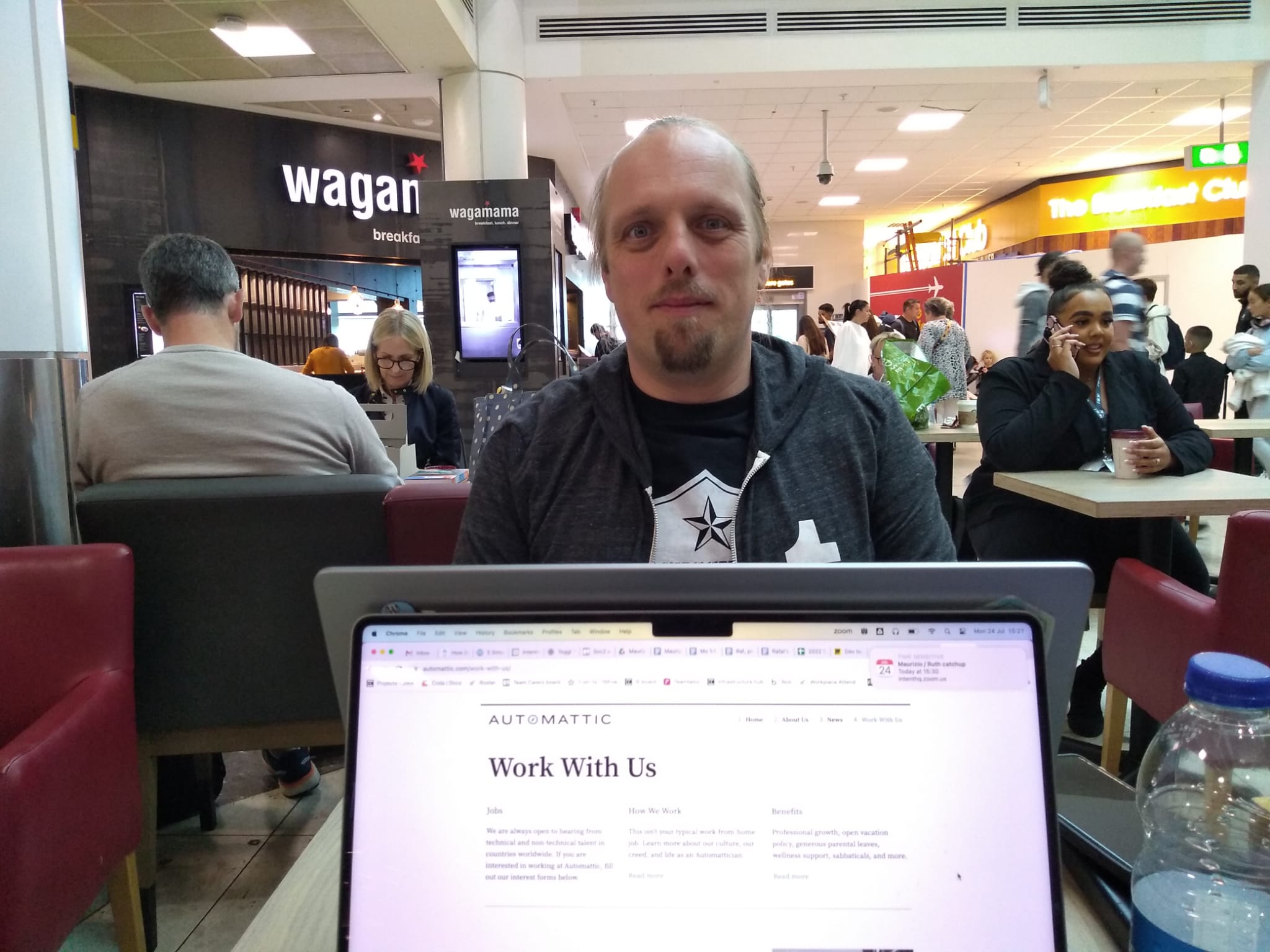 A laptop screen shows Automattic's "Work With Us" web page. Beyond it, in an airport departure lounge (with diners of Wagamama and The Breakfast Club in the background), Dan sits at another laptop, wearing a black "Accessibility Woke Platoon" t-shirt and grey Tumblr hoodie.