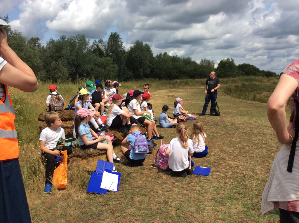A group of schoolchildren in a mixture of white and blue shirts, and with most wearing sunhats, sit on a pile of rocks alongside a ring ditch and listen intently to Dan.
