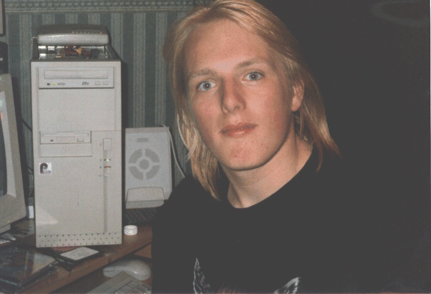 Photograph of Dan in his mid-teens, with shoulder-length bleached-blonde hair and wearing a t-shirt with a picture of a snarling wolf, sits in front of a running PC (with its beige case open) on which an external modem is precariously balanced.