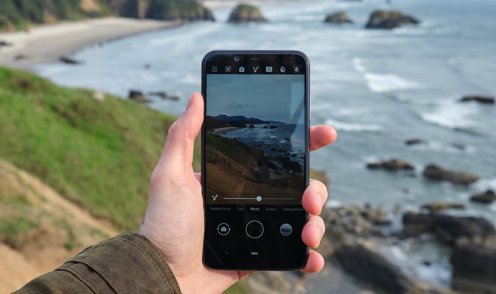 Photo showing a mobile phone, held in a hand, being used to take a photograph of a rugged coastline landscape.
