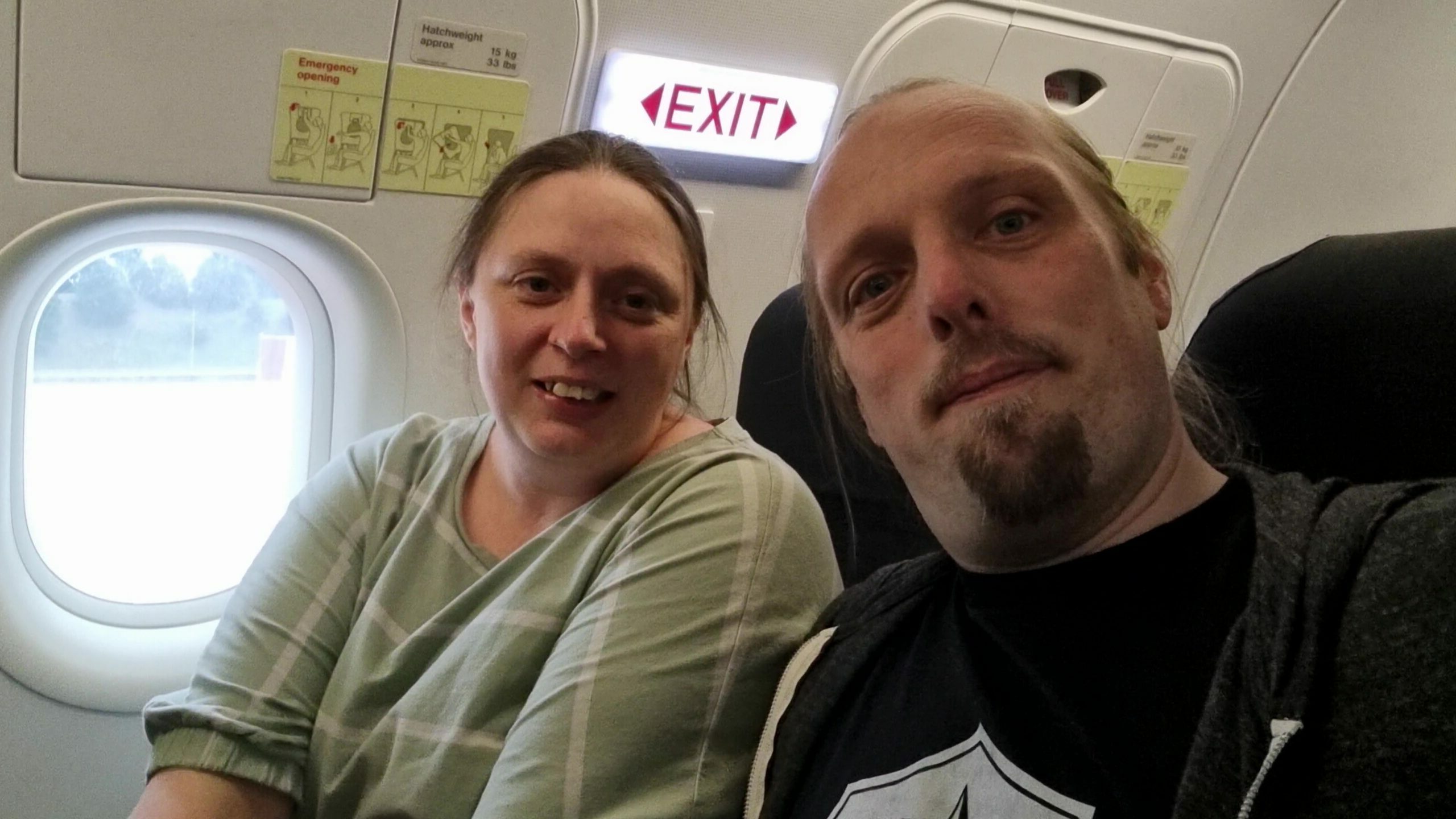 Ruth, wearing a green top with white stripes, sits alongside Dan, wearing a black t-shirt and grey hoodie, by the wingside emergency exits in an aeroplane.