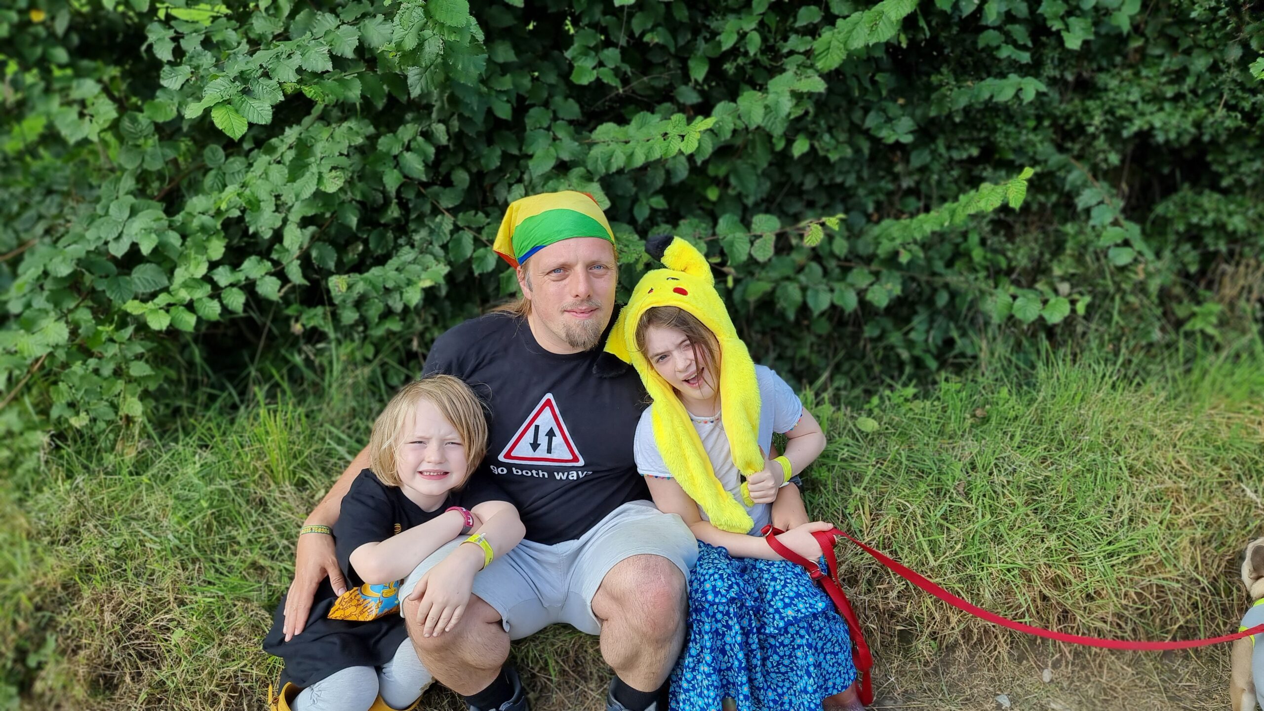 Photograph showing Dan, wearing a "go both ways" t-shirt and a rainbow-striped bandana and grey shorts, sits between a 6-year-old boy and a 9-year-old girl (the girl is wearing a Pikachu hat).