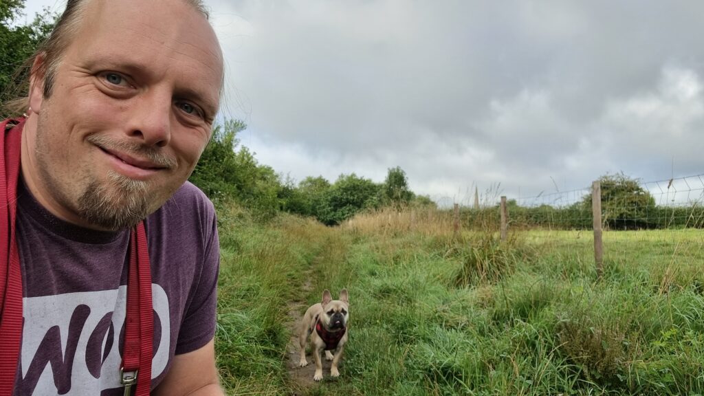 Dan, wearing a purple "Woo" t-shirt, crouches by Demmy, a champagne-coloured French Bulldog, in a path through the long grass between a wood and a field. In the distance, the church spire of St Peter & St Paul at Church Hanborough can be seen protuding above the treeline.