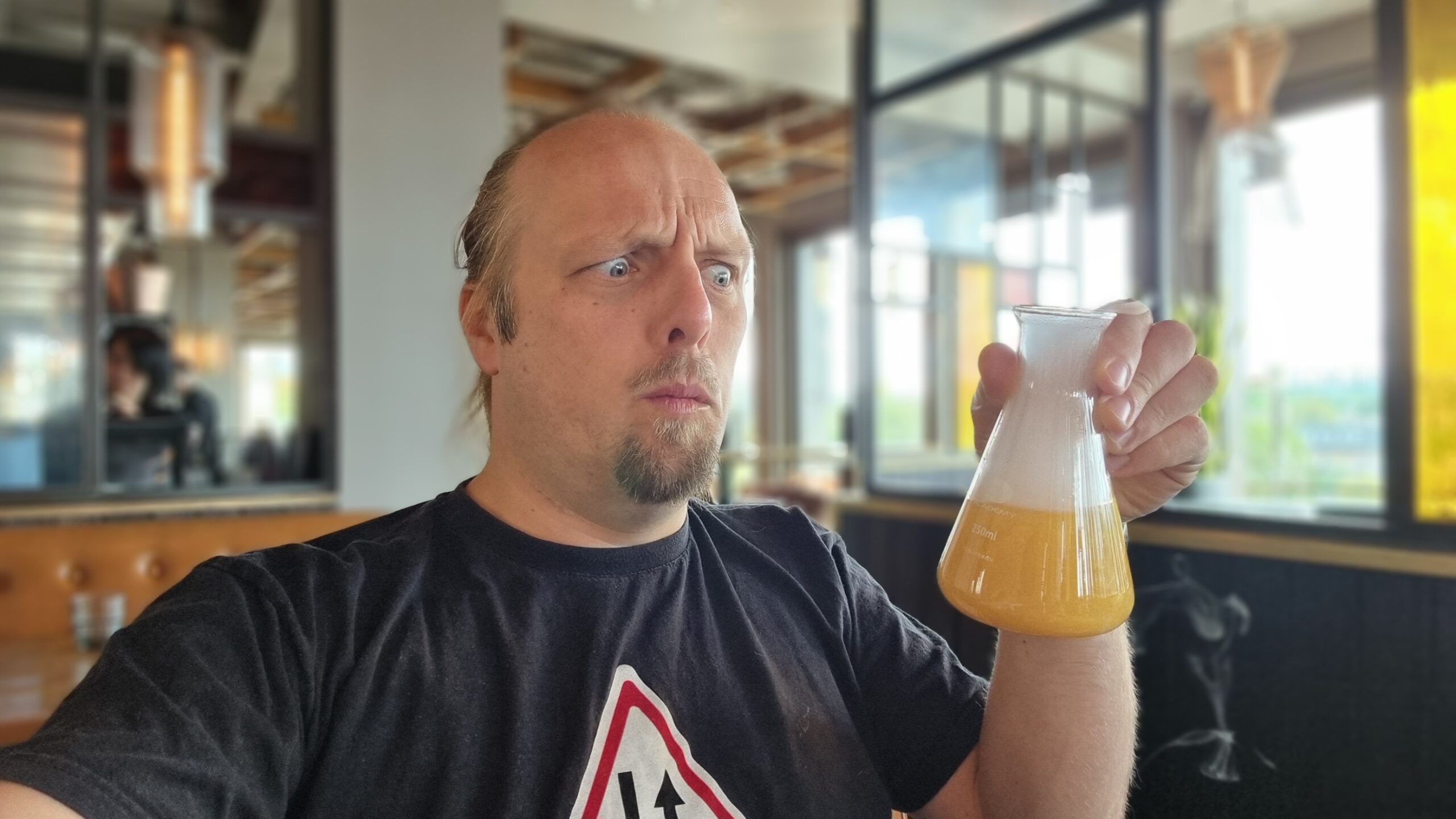 Selfie photograph of Dan, in a bar with a rooftop view of daylight out the windows in the background, looks concerned as he stares at the a frothy, bubbling flask of yellow liquid he's holding.