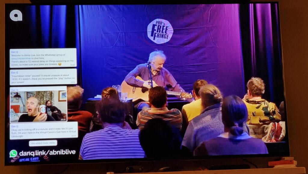 Phootgraph of a wall-mounted television screen. On the screen, comedian Peter Buckley Hill sits with his guitar on his lap in front of an audience: the "PBH's Free Fringe" logo is on the curtain behind him. On the left of the screen a series of WhatsApp messages appear, including one showing a photo of Dan holding a can of Old Speckled Hen beer.