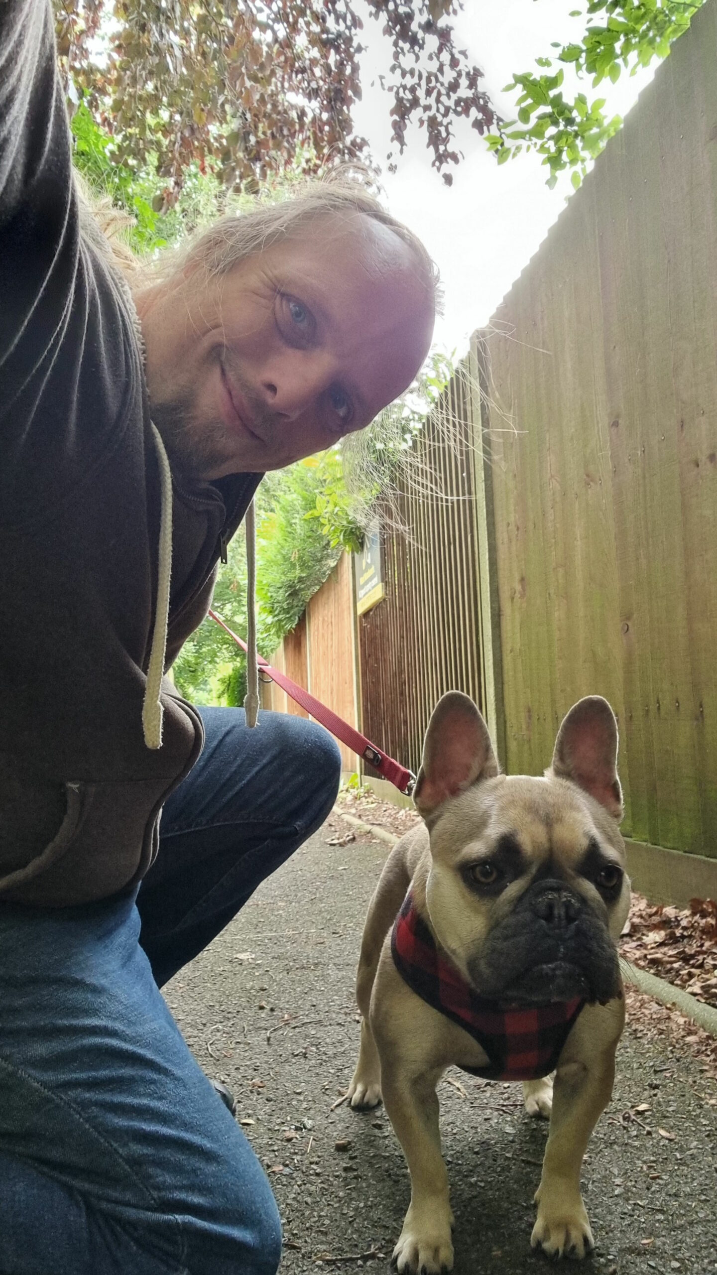 Dan, wearing blue jeans and a grey hoodie, kneels alongside Demmy, a French Bulldog, on a dirt path between a forest and the wooden fences at the edge of some gardens.