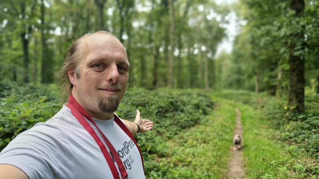 Dan, wearing a white t-shirt and with a red dog lead hanging over his shoulder, stands in a forest, gesturing down a path to a small French Bulldog following him.