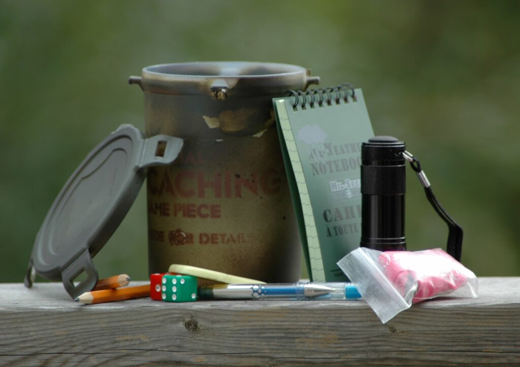 Photograph showing a medium-sized geocache container with its contents laid-out around it: various pieces of swag for trade, plus a notebook.