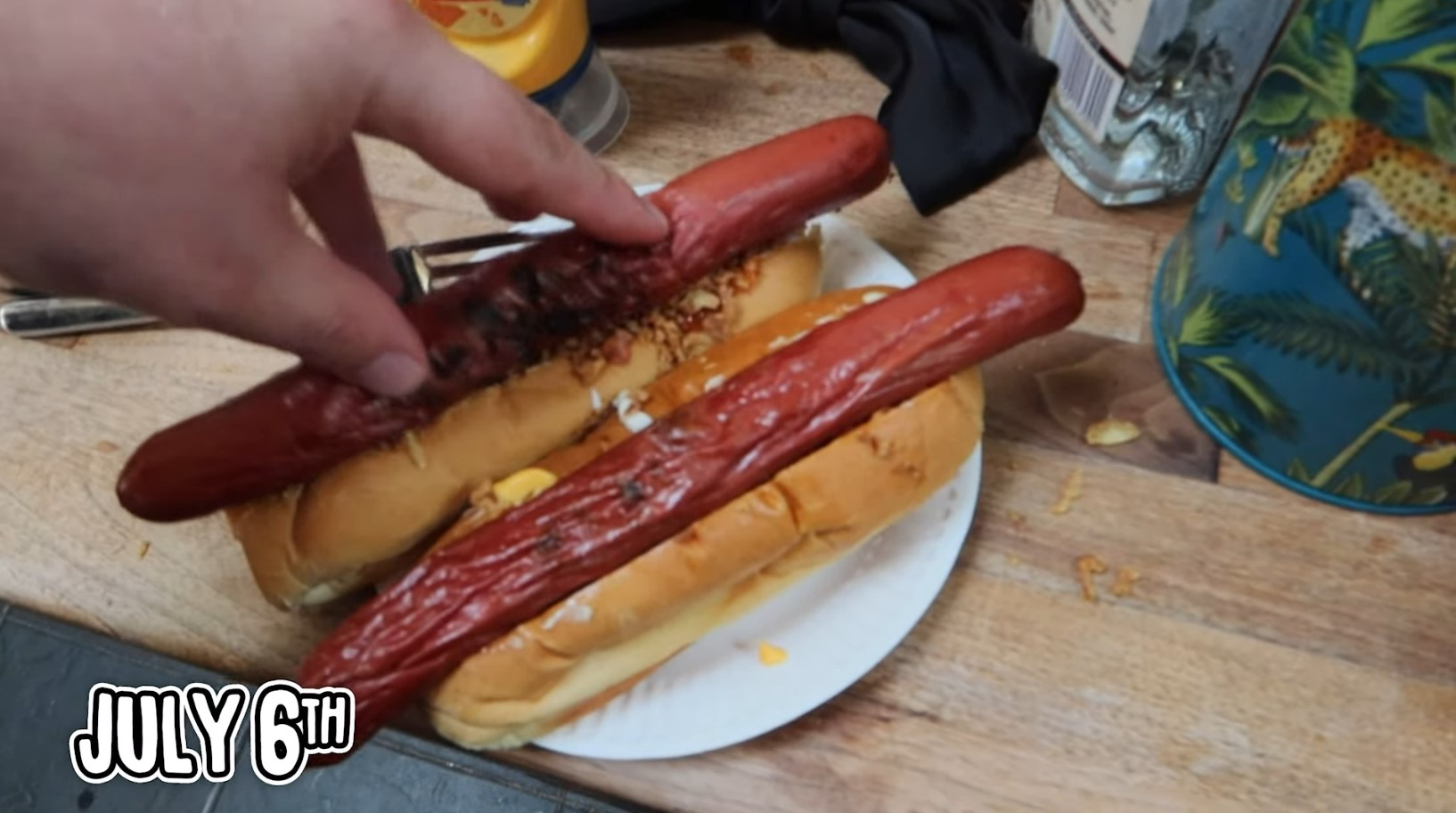 Frame from a video showing TomSka's hand pushing a hot dog sausage into a bun, on top of fried onions and sauces. The picture is captioned "July 6th".