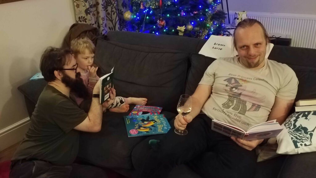 Dan, wearing a Princess Twilight Sparkle / Frank Herbert's Dune crossover fan art t-shirt, sits on a grey sofa in front of a lit Christmas tree, holding a glass of wine. At the other end of the sofa JTA, a white man with a thick beard and glasses, reads to a (tired-looking) young boy. All three are surrounded by books.