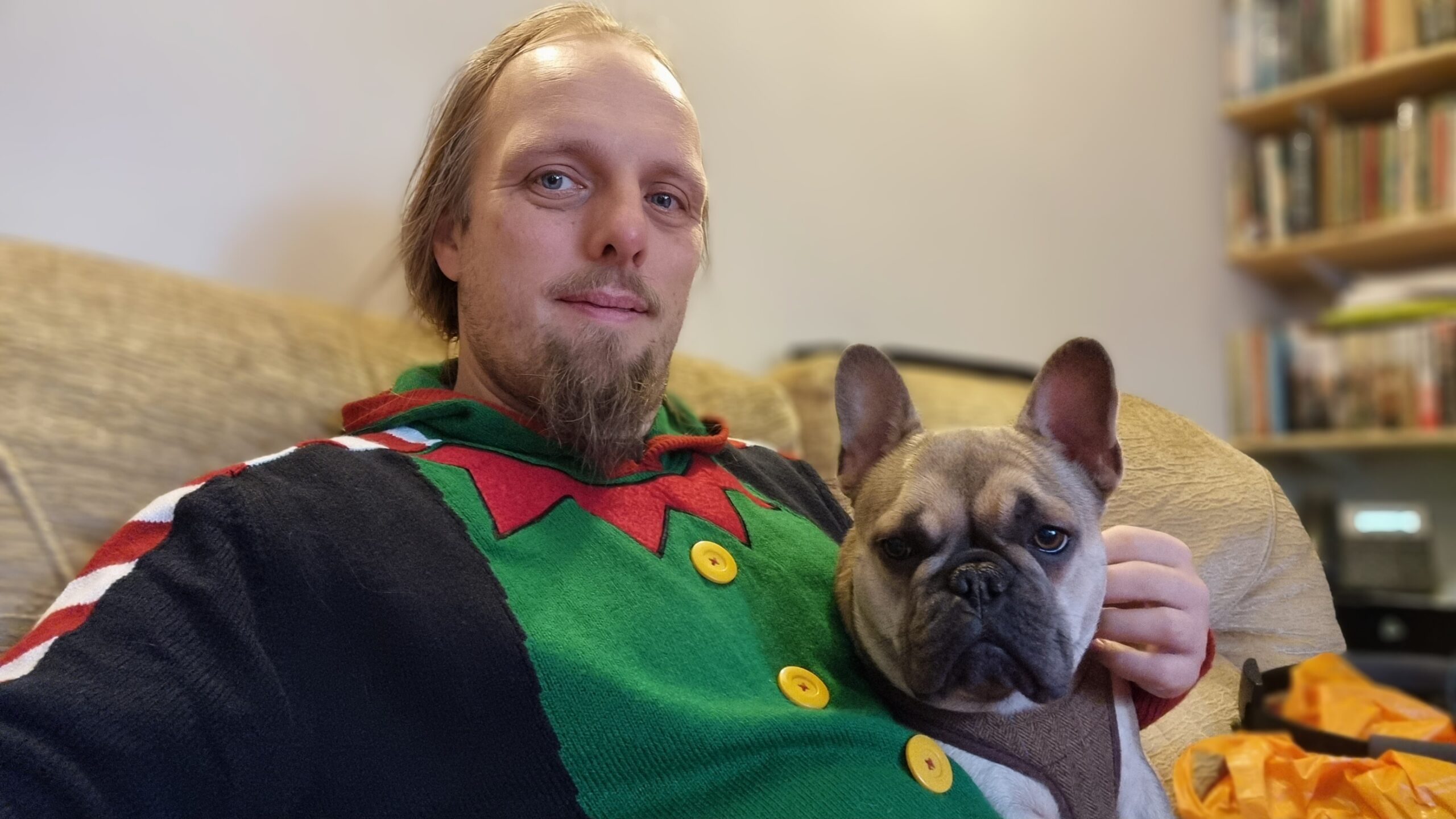 Dan, wearing an "elf costume" Christmas jumper, looks into the camera while cuddling a French Bulldog. The pair are sitting on a beige sofa.