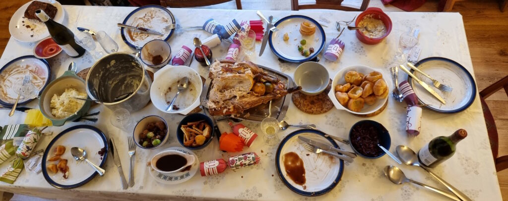 Top-down view of a dining table set with a Christmas-themed tablecloth. The meal has concluded and the seats have been vacated, but large amounts of food - most of a turkey, half a nutloaf, lots of mashed potato, several sprouts, stuffing balls, and chestnuts, some roast potatoes and parsnips, an entire boat full of gravy, and almost a dozen Yorkshire puddings - are still set out.
