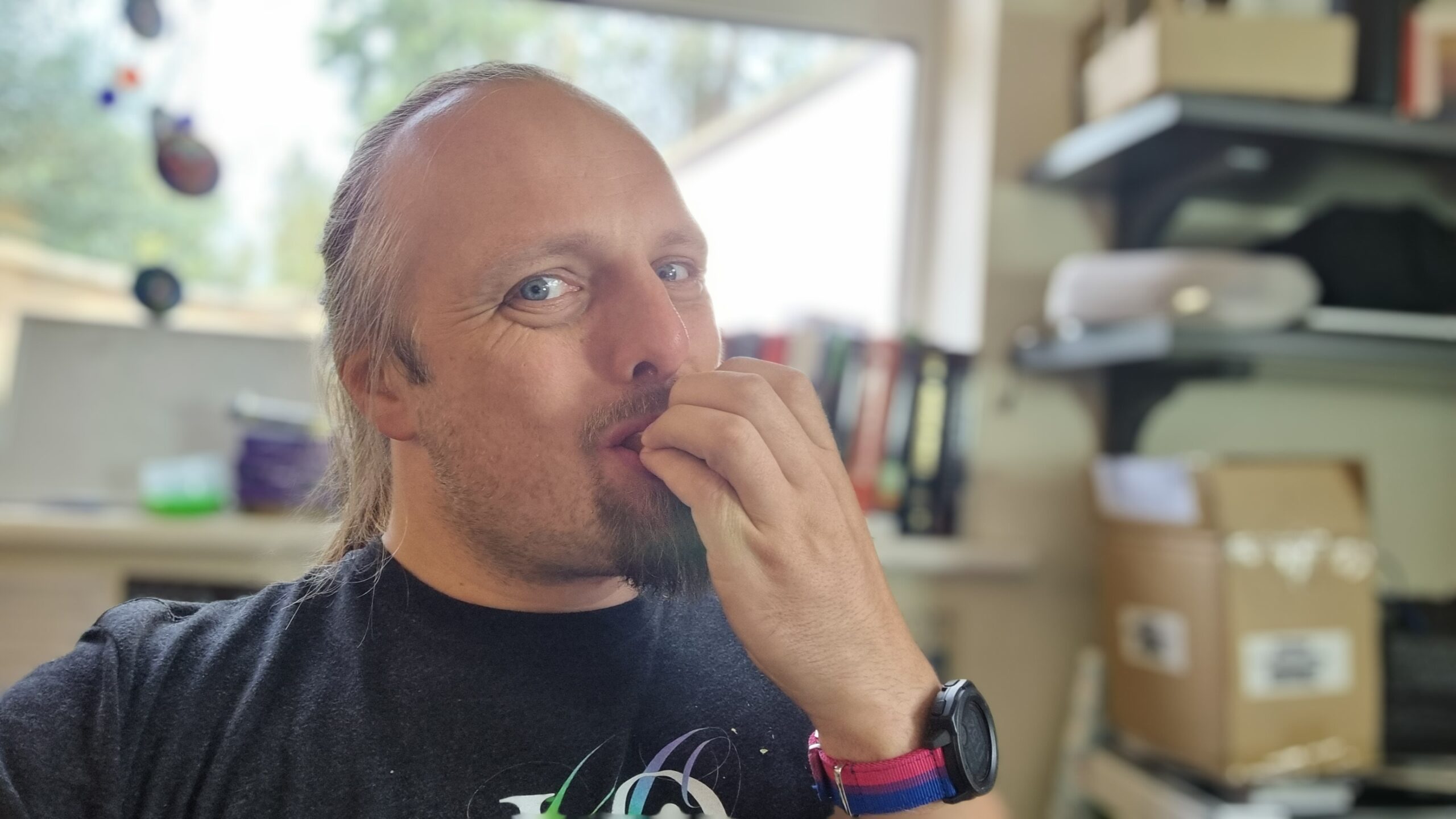 Dan - a white man with long hair tied back and a goatee-style beard - bites into something concealed by his hand (but implied to be a crisp sandwich), which is at his mouth. He's wearing a black t-shirt on which the top part of a swirling rainbow can be made out, and a watch with a "bi pride"-flag coloured watch strap. A window and a cluttered office space are out-of-focus behind him.