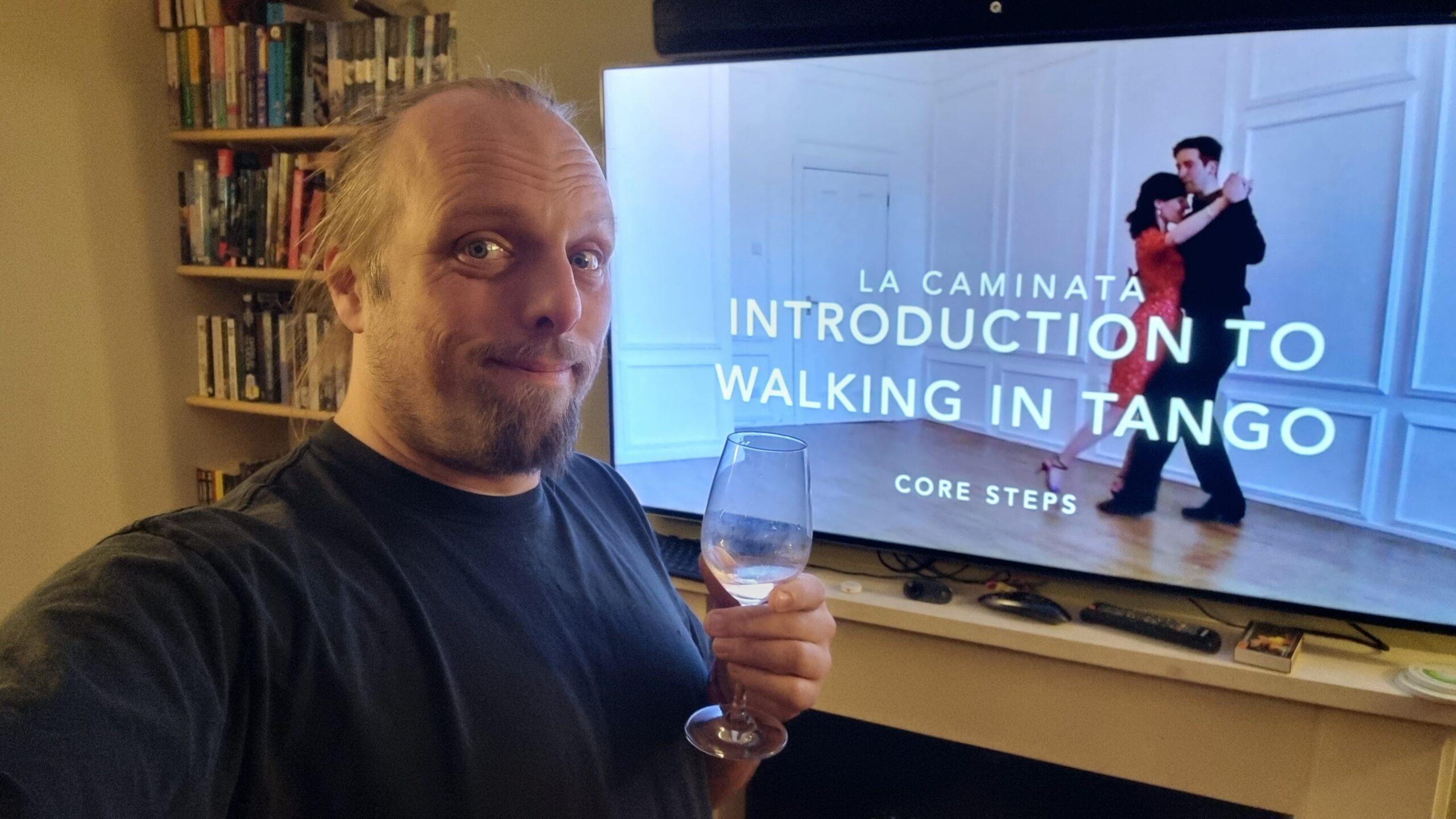 Dan, wearing a black t-shirt and holding a glass of wine, looks sceptically at the camera as he stands in front of a television screen showing a couple dancing, with the title frame "La Caminata: Introduction to Walking in Tango (Core Steps)".