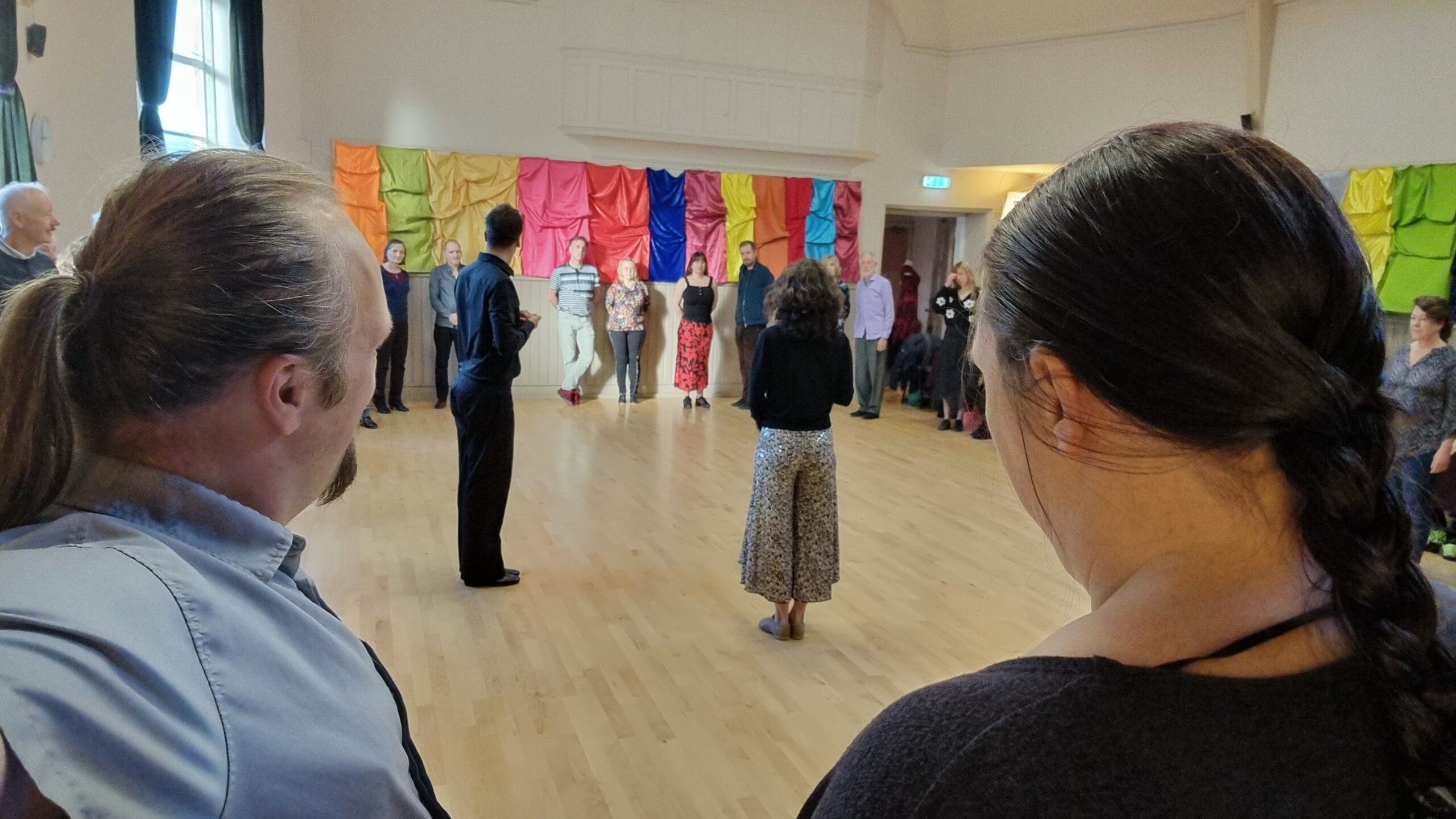 In a church hall, its walls decorated with colourful cloths, Dan and Ruth stand in a large circle of people, watching a man and a woman preparing to demonstrate some tango steps.