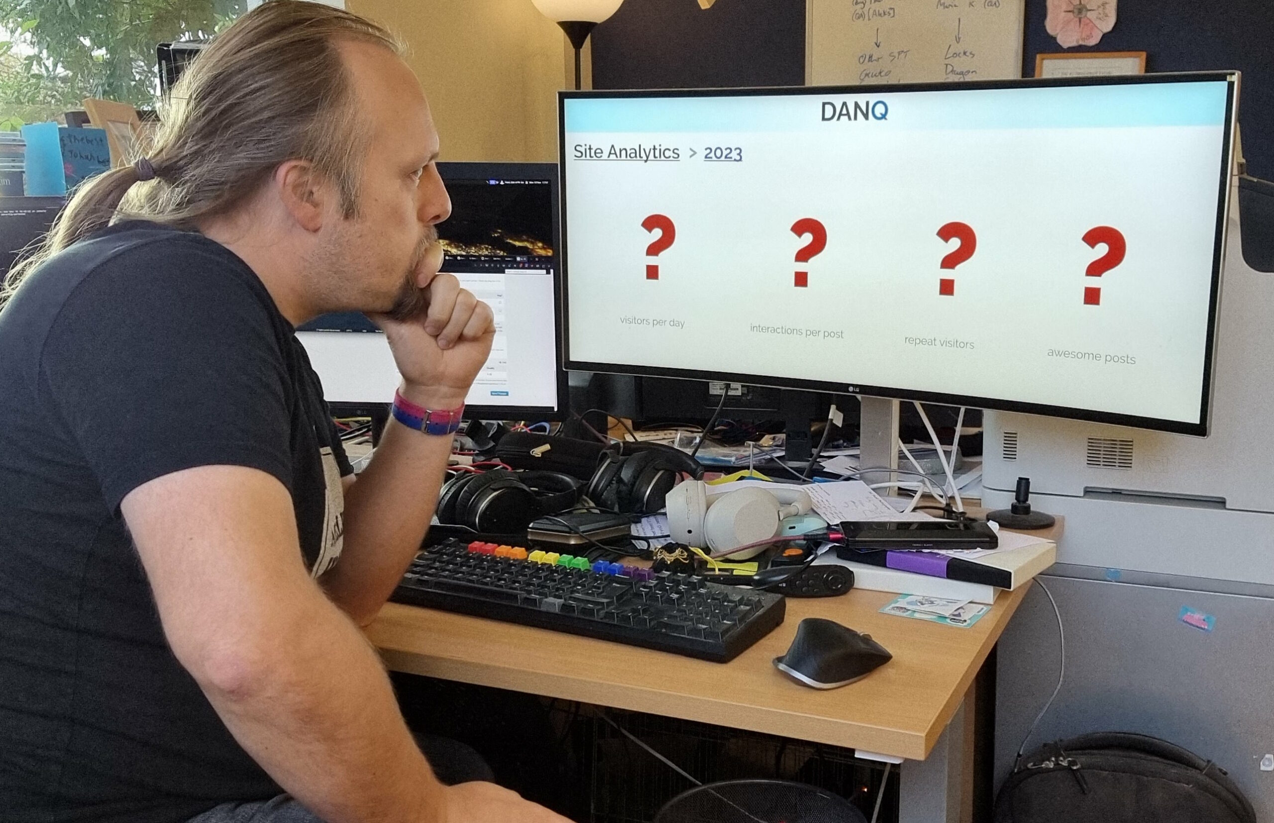 Dan, wearing a black t-shirt and jeans, sits hunched over a keyboard with Pride-coloured keys, looking thoughtfully at a widescreen monitor. On the monitor is a mocked-up screenshot showing site analytics for DanQ.me, but with question marks for every datapoint.