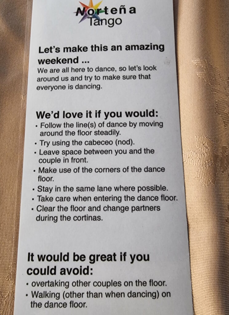 Photograph of a small laminated instruction sheet on a golden tablecloth. Titled "Norteña Tango", it reads: Let's make this an amazing weekend. We are all here to dance, so let's look around us and try to make sure that everyone is dancing. We'd love it if you would follow the lines of dance by moving around the floor steadily, try using the cabeceo, leave space between you and the couple in front, make use of the corners of the dance floor, stay in the same lane where possible, take care when entering the dance floor, clear the floor and change partners during the cortinas. It would be great if you could avoid overtaking other couples on the floor, walking (other than when dancing) on the floor.