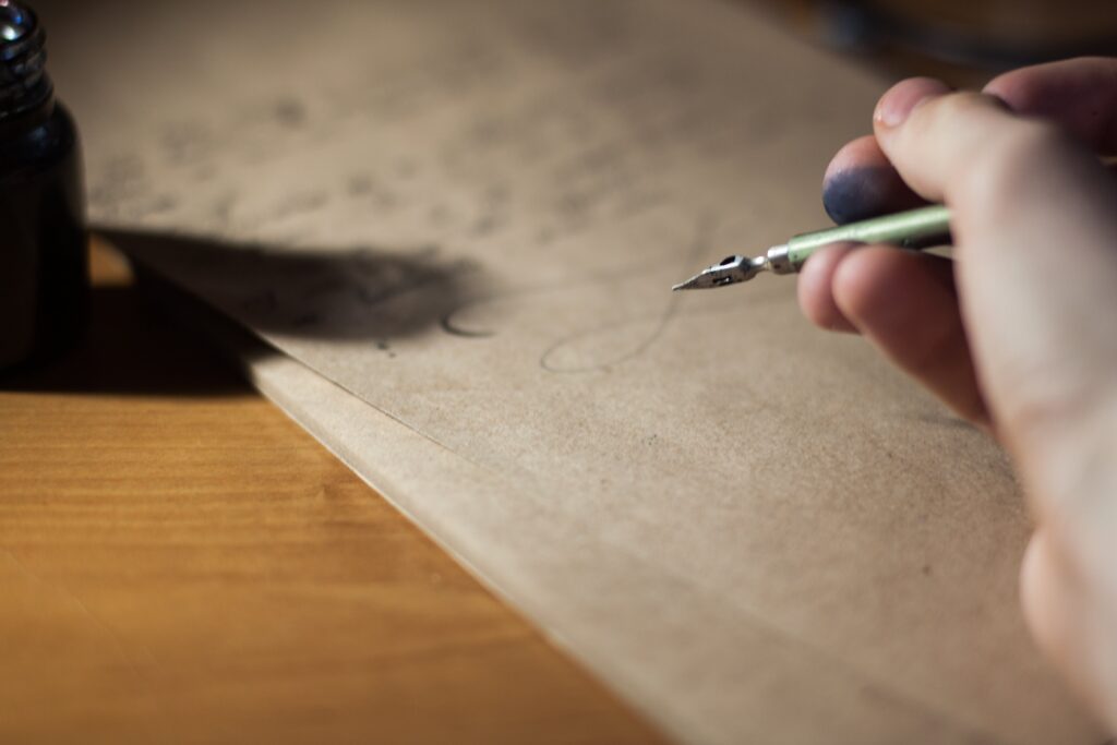 Traditional inkwell and pen, the latter held in an inkstained finger grip, being used to write a letter on unbleached paper atop a wooden desk.