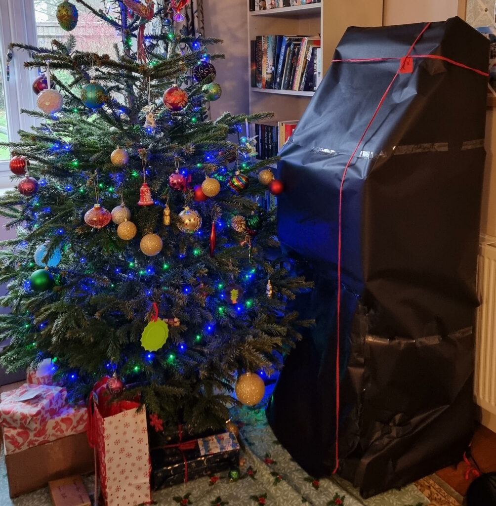 A large, arcade-cabinet-shaped present, wrapped in black paper and a red ribbon, stands alongside a Christmas tree.