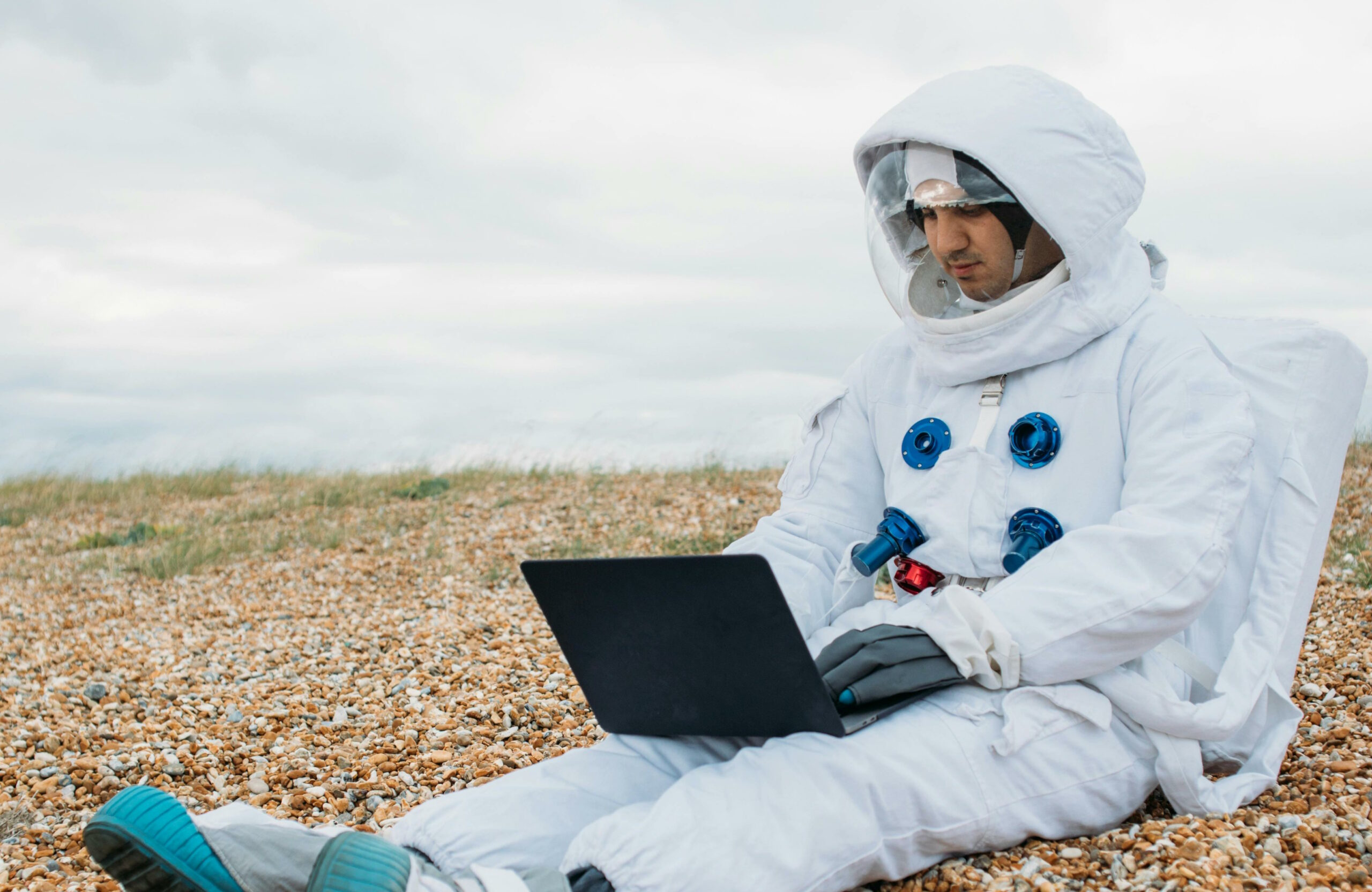 A white man wearing a spacesuit sits on a pebble beach using a laptop.