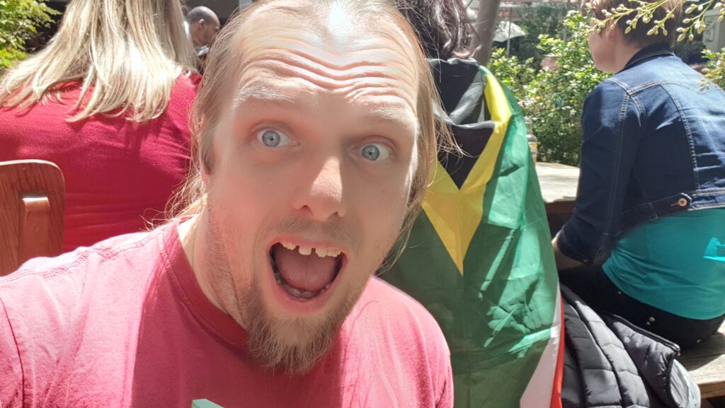 Dan, wearing a red t-shirt and in bright sunshine, pretends (badly) to be shouting excitedly. In the background, a group of people are watching a televised rugby match; one is wearing a South African flag as a cape.